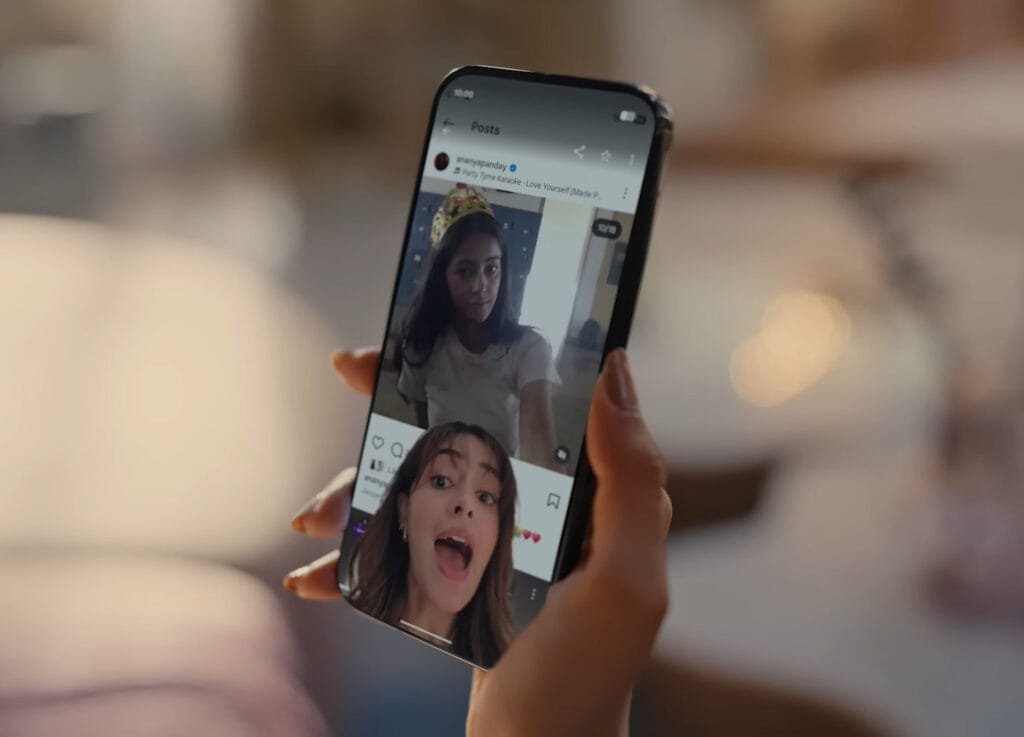 Pessoa segurando smartphone exibindo o recurso Screen Reactions com gravação simultânea da tela e da câmera frontal, mostrando duas pessoas em vídeo de reação.